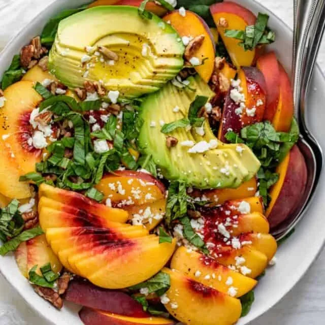 Peach salad with avocado slices, spinach, feta, and pecans in a white bowl