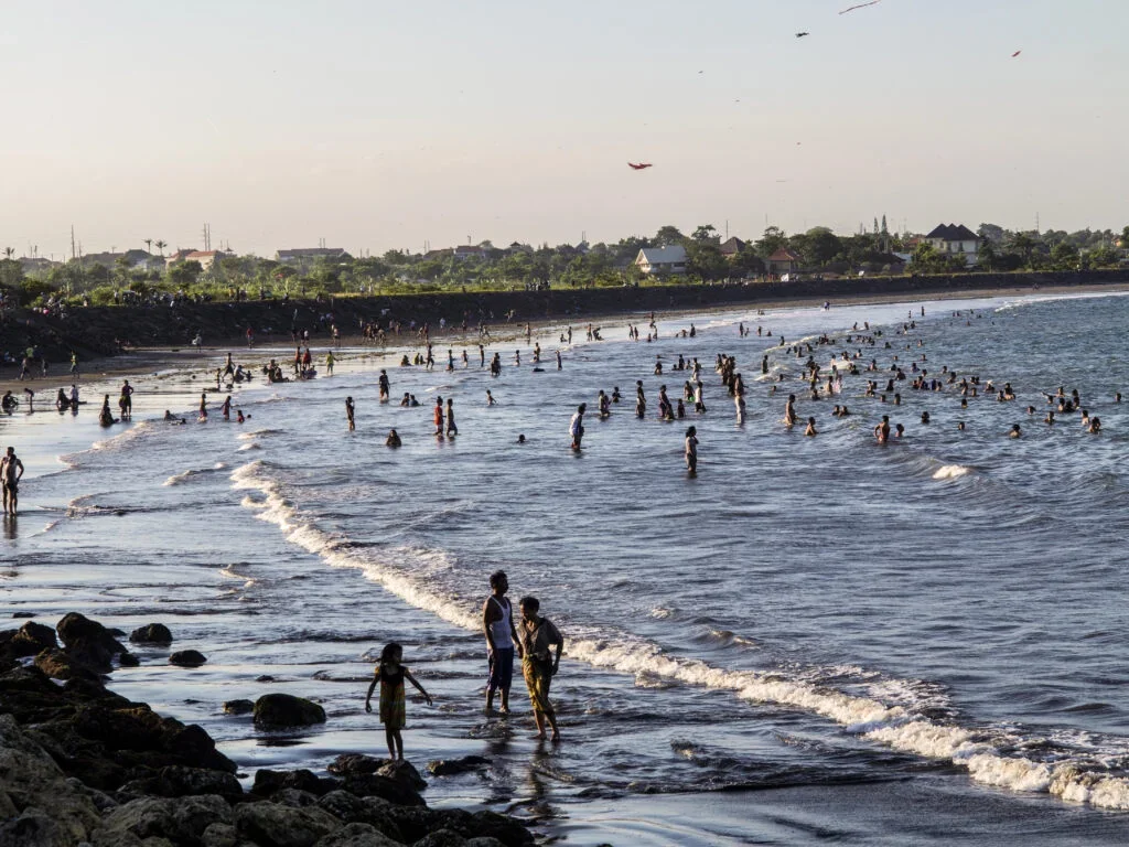 Crowded Bali beach with dark volcanic sand and tourists under palm trees