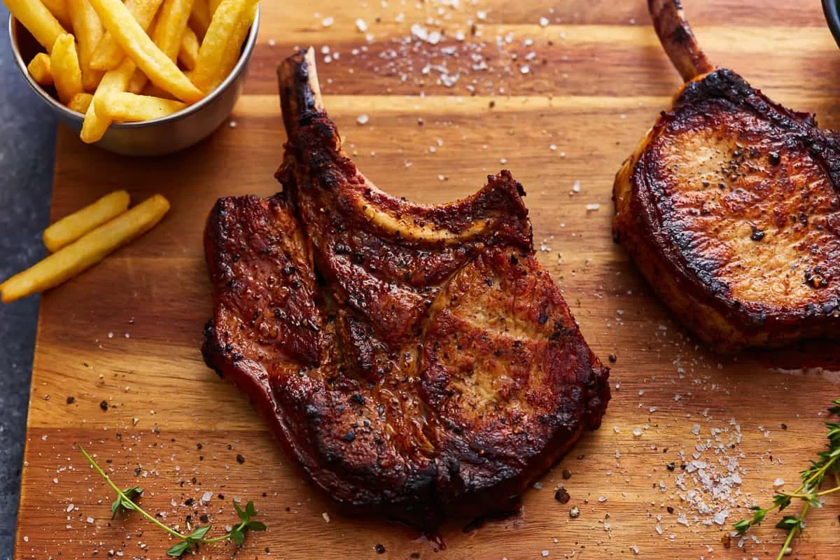 Cast Iron Pork Chops Recipe - The Cookie Rookie® 1 raw bone-in pork chops with fresh herbs, garlic, butter, and oil on a wooden board