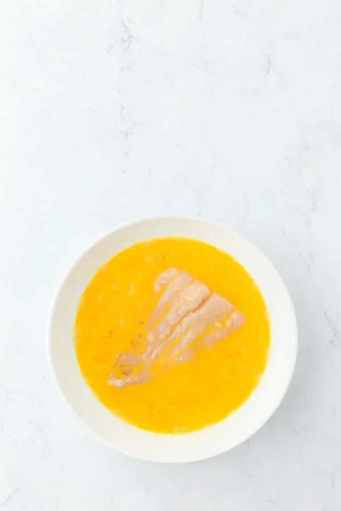 Authentic Mississippi Fried Catfish Recipe That's Crispy & Tender 4 raw catfish fillet being dipped in a bowl of egg wash