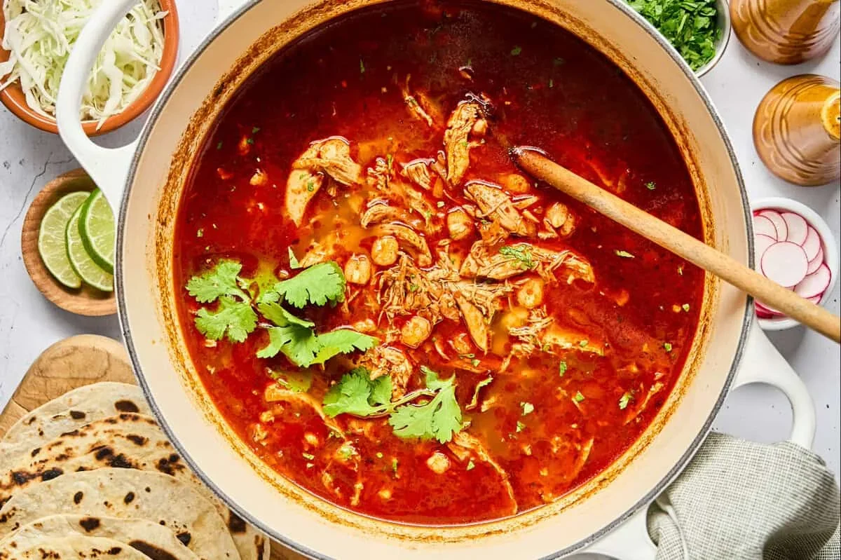 A bowl of red chicken pozole rojo with hominy, topped with cabbage, radish, and lime